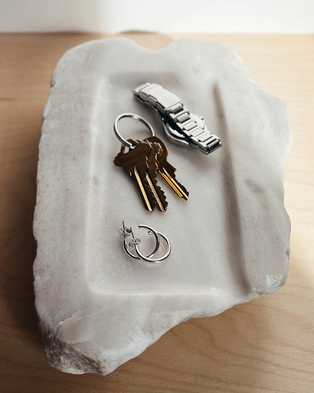 Front-on: organic-shaped off-white alabaster tray holding earrings, keys and watch.